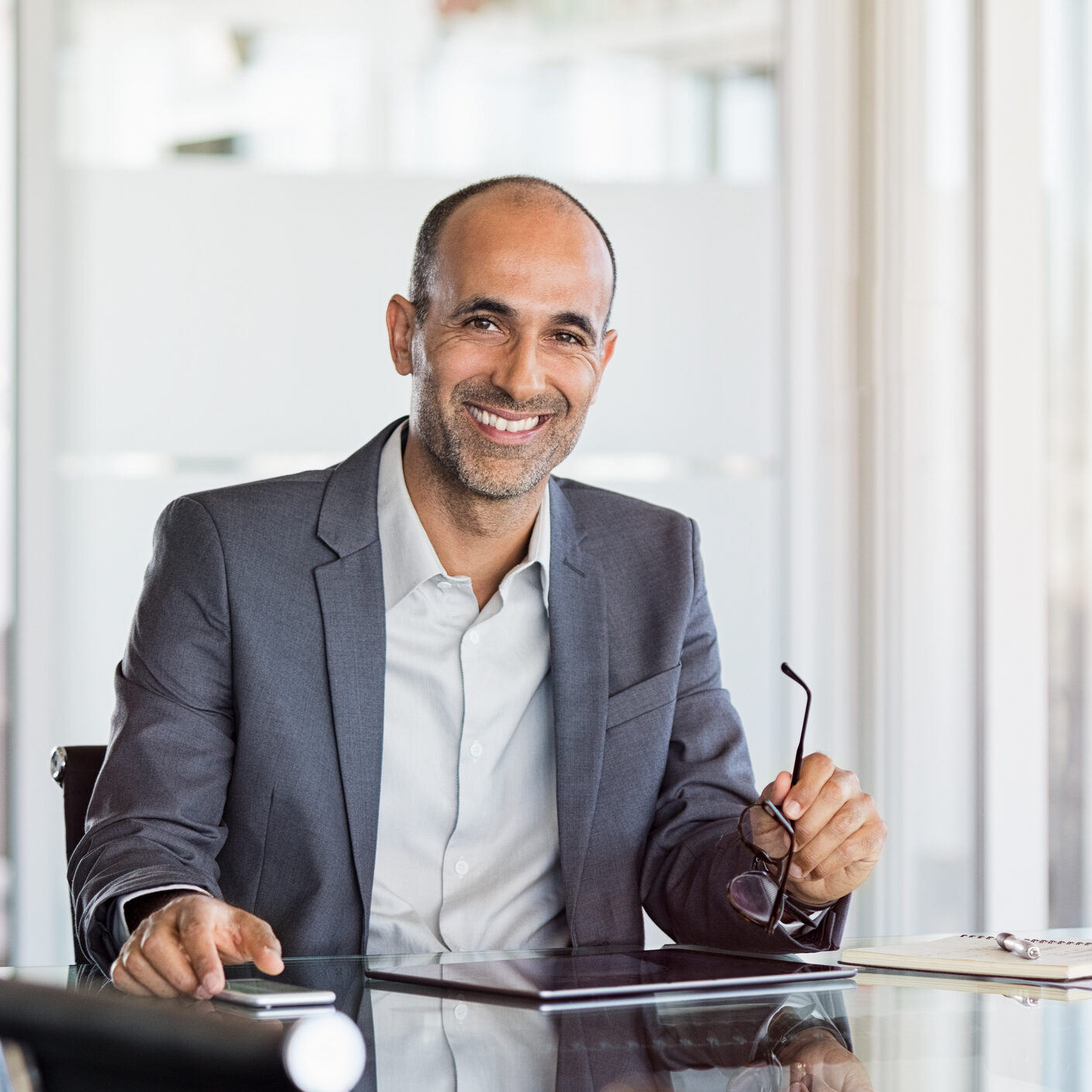 Happy mature business man holding spectacles in modern office. Successful senior businessman in formals sitting in meeting room with phone and tablet. Smiling man in suit in a elegant office.